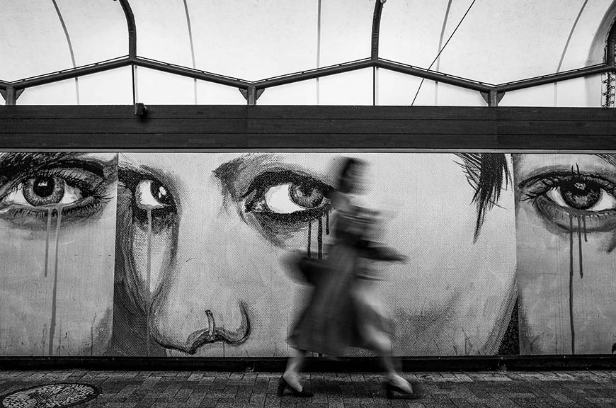 A black and white photo of a woman walking past a mural.