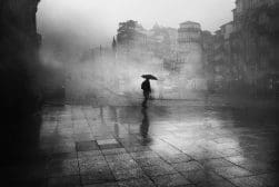 A black and white photo of a person walking down a street with an umbrella.