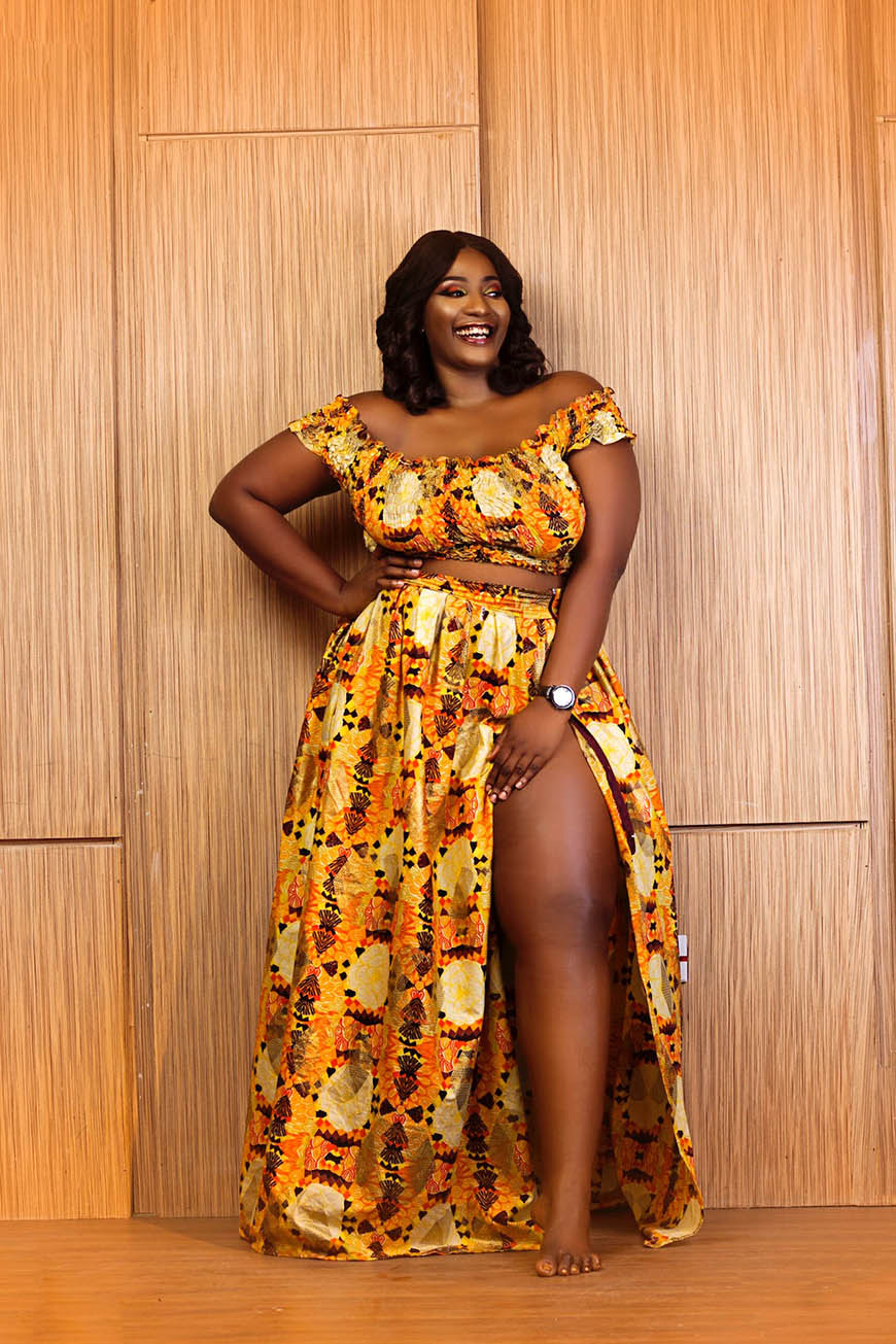 A woman in an african print dress posing in front of a wooden wall.