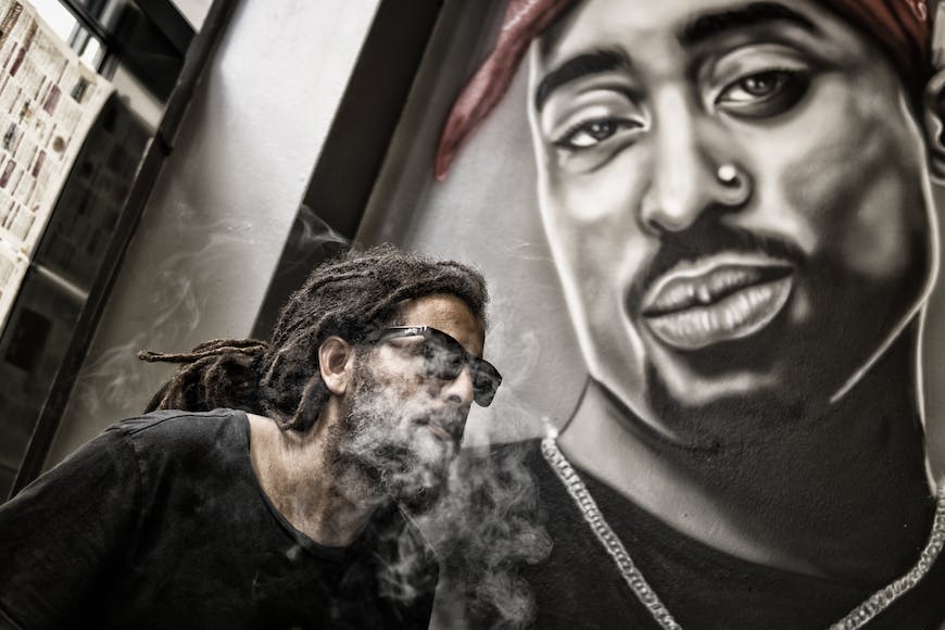 A man with dreadlocks smoking a cigarette in front of a mural.