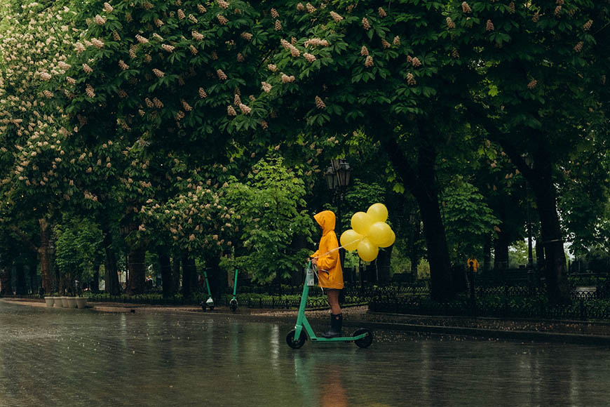A person riding a scooter in the rain with yellow balloons.