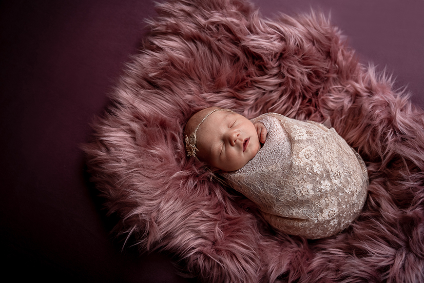 A newborn baby wrapped in a fur blanket on a pink background.