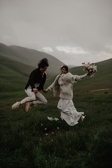 A bride and groom jumping in the air in a field.
