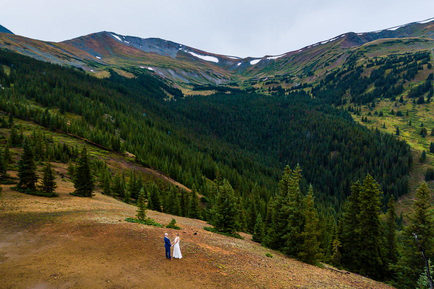 A bride and groom standing on top of a hill in the mountains.