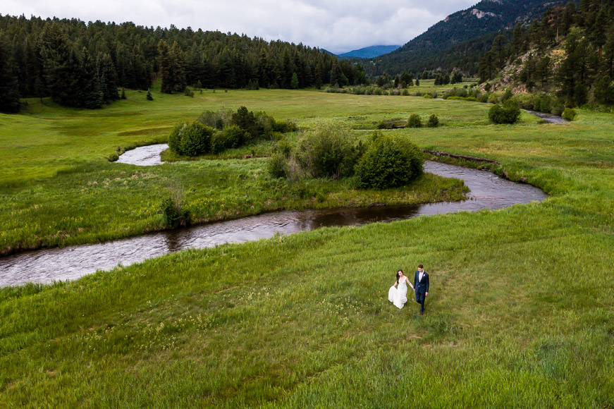 A bride and groom walking along a river in a grassy meadow.