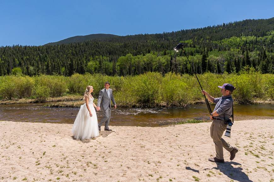 A bride and groom standing on a beach with a camera.