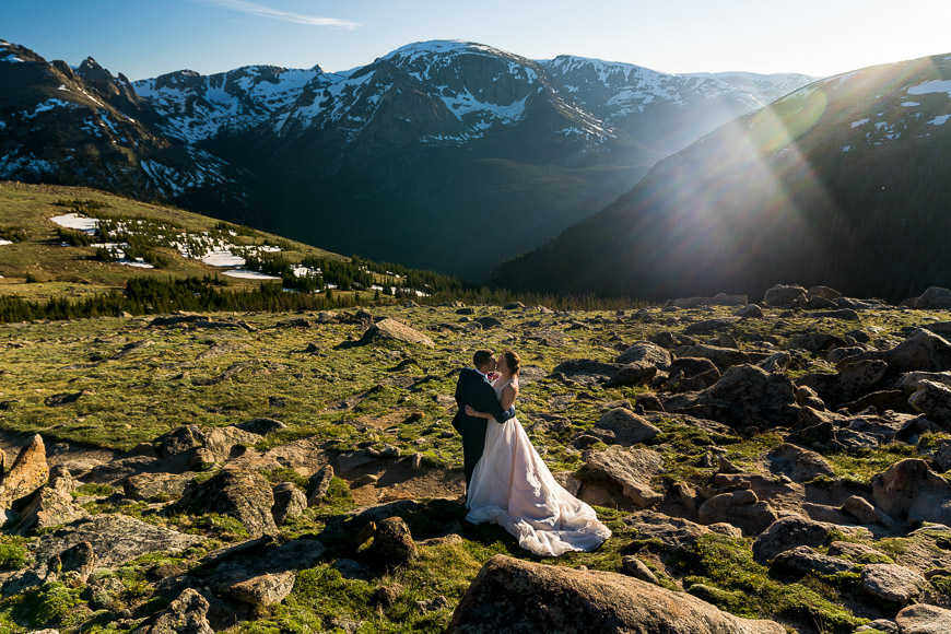 A bride and groom standing on top of a rocky mountain with the sun shining down on them.