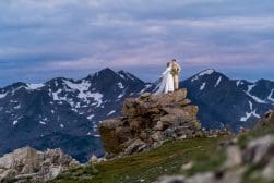 A bride and groom standing on top of a rock in the mountains.