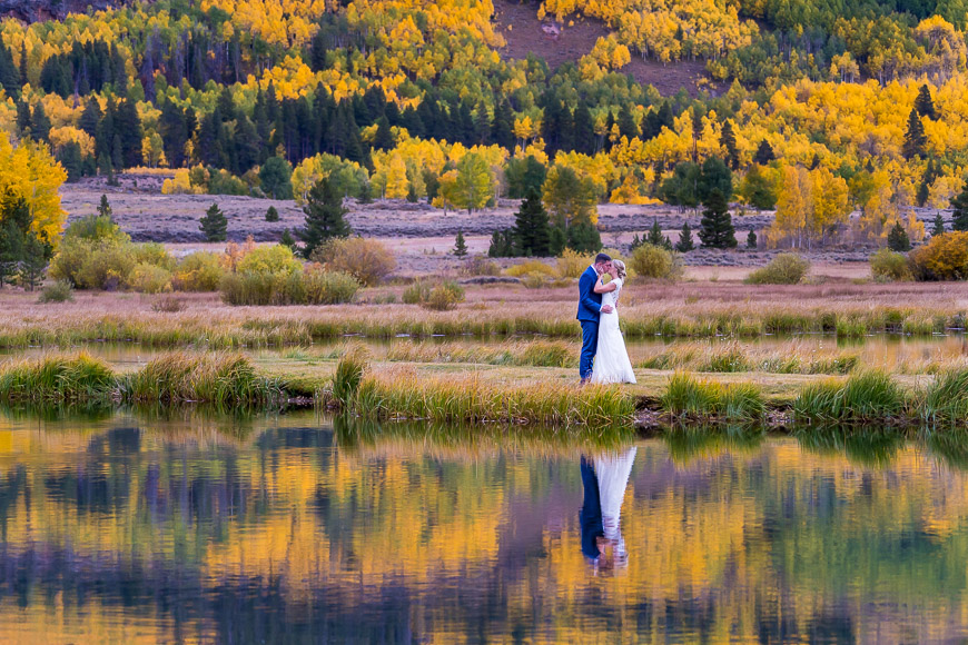 A bride and groom standing in front of a pond with autumn trees in the background.