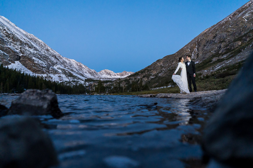 A bride and groom standing in front of a mountain lake at dusk.