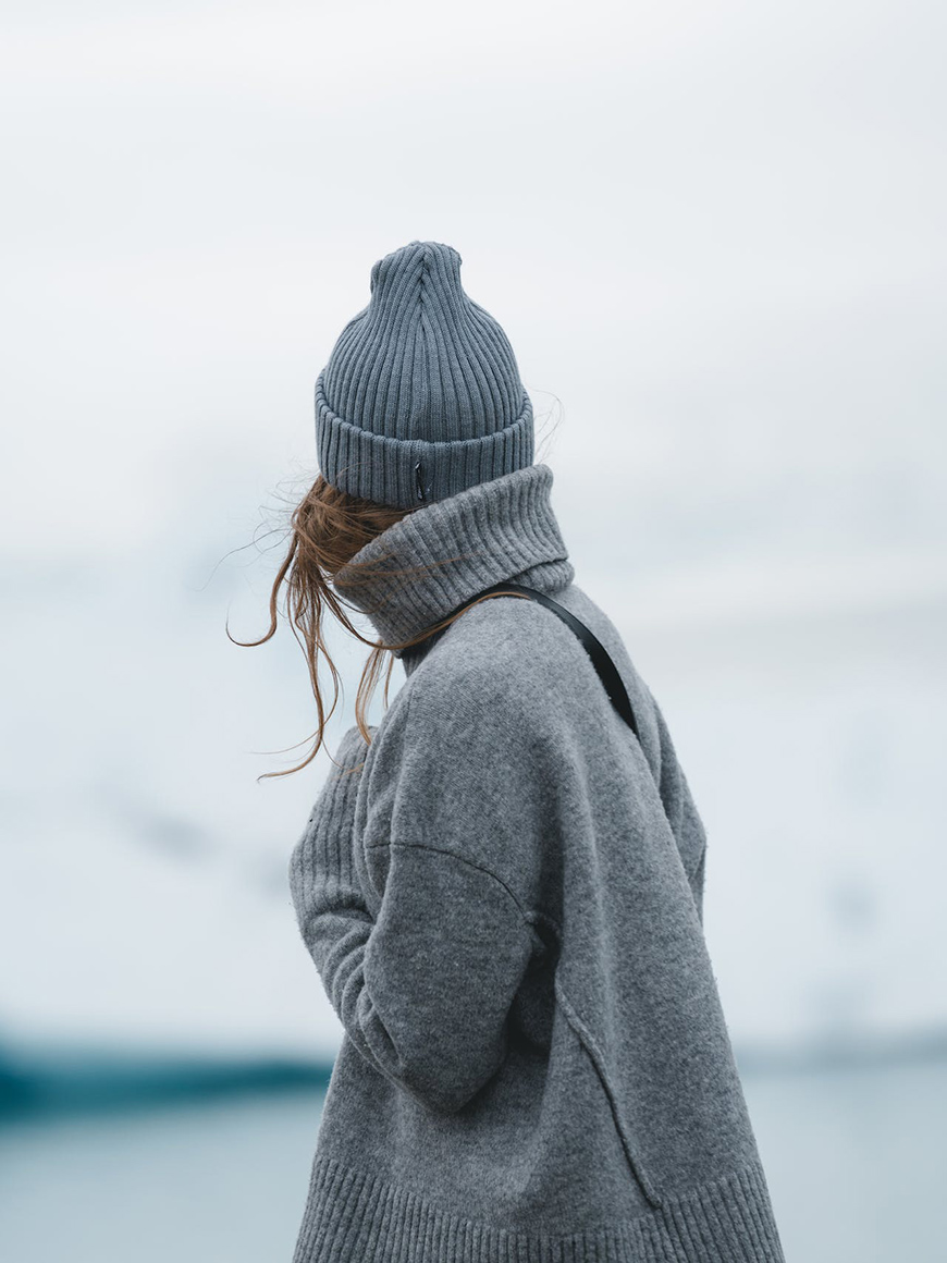 A woman wearing a grey sweater and a beanie in front of an iceberg.