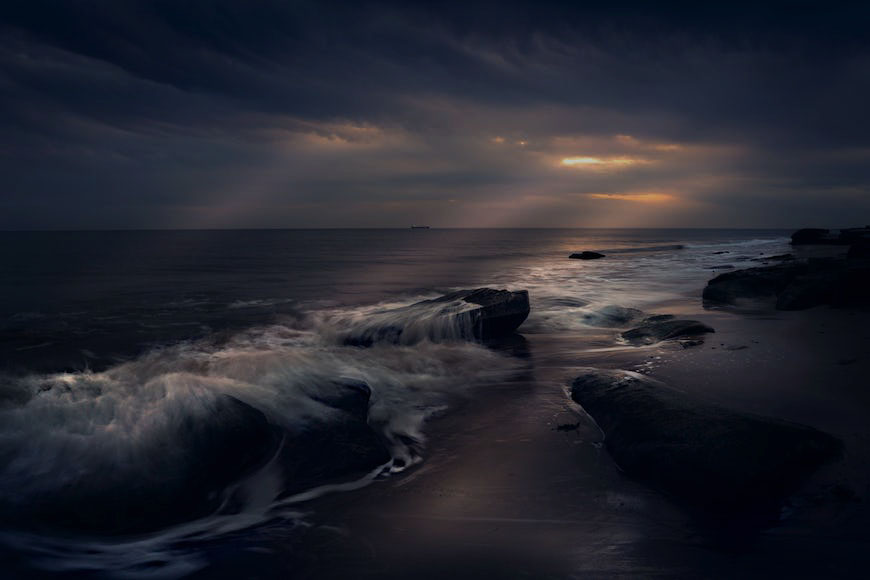 A dark stormy sky over a beach with rocks and waves.