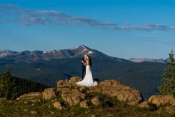A bride and groom standing on top of a rock with mountains in the background.