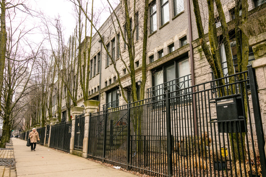 A person walking down a sidewalk beside a black metal fence, with a row of modern townhouses lined with bare trees.