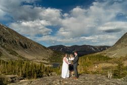 A bride and groom standing on top of a rock in the mountains.