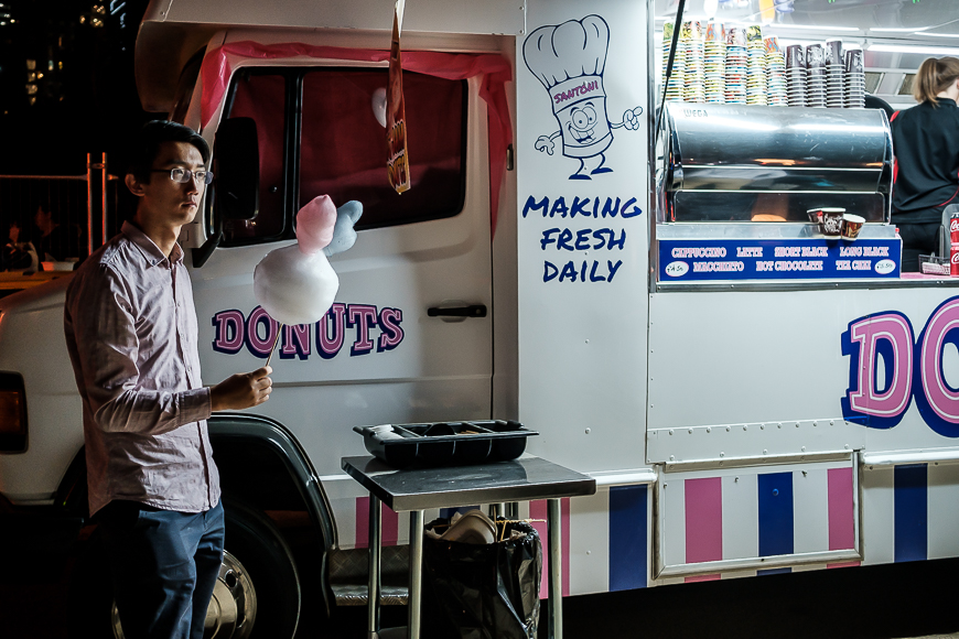 Man holding cotton candy stands beside a donut food truck at night.