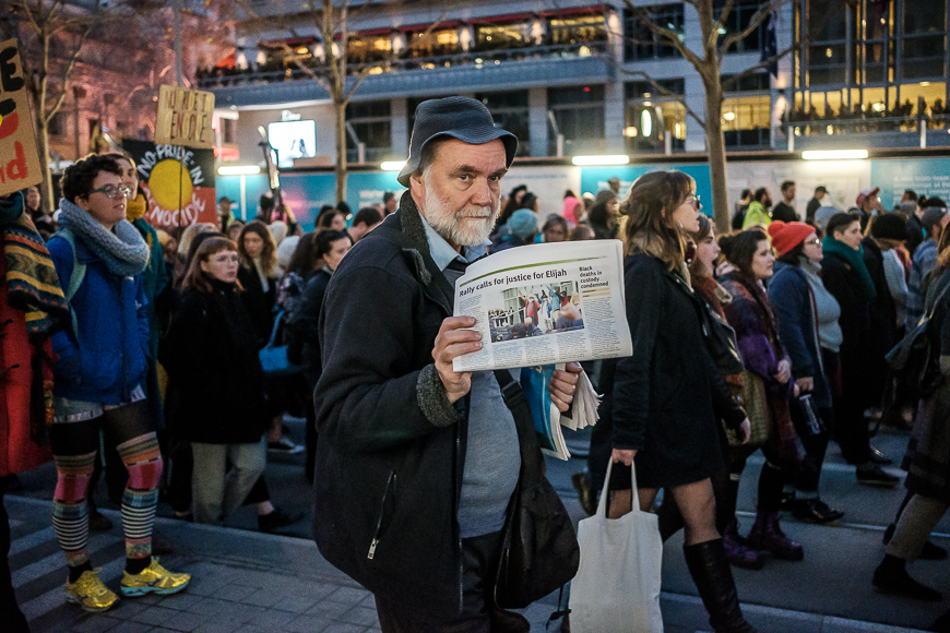 Man holding a newspaper stands out from a crowd of protesters in an urban setting.