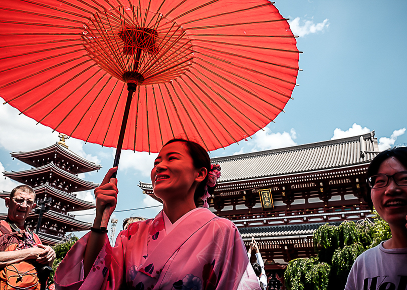 A woman in a pink kimono holding a red parasol with a pagoda and people in the background.