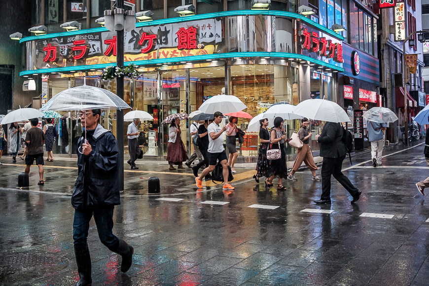 Pedestrians with umbrellas walking on a rainy city street with neon signs.