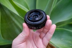 A camera lens held in a person's hand against a green plant background.