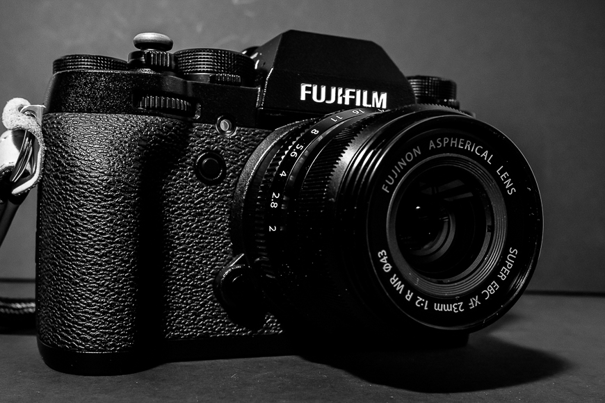 Black and white photo of a fujifilm camera with a prime lens, resting on a textured surface.