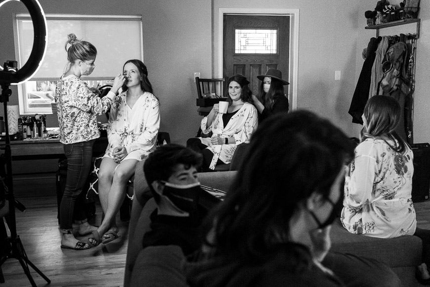 A makeup artist applies cosmetics to a seated woman in a robe while others wait and watch in a dressing room.