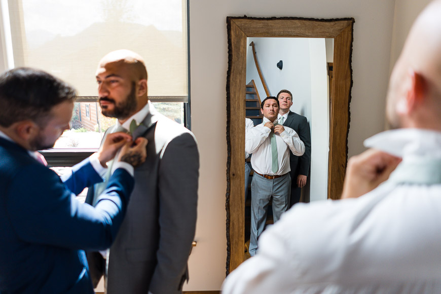 Two men adjusting ties in a mirror while a third assists one of them.