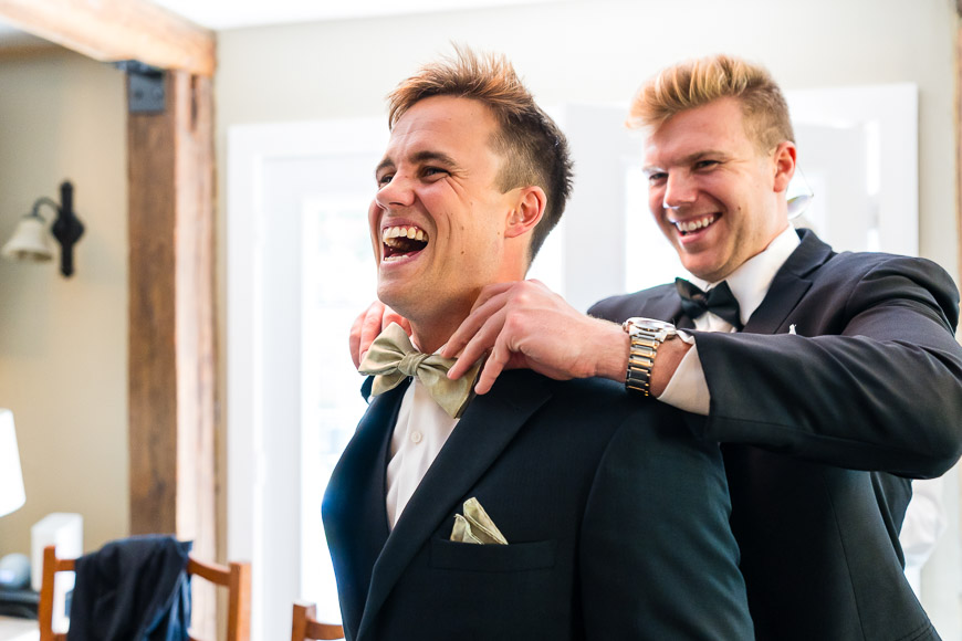 Two men in formal attire sharing a joyful moment, with one adjusting the other's bow tie.