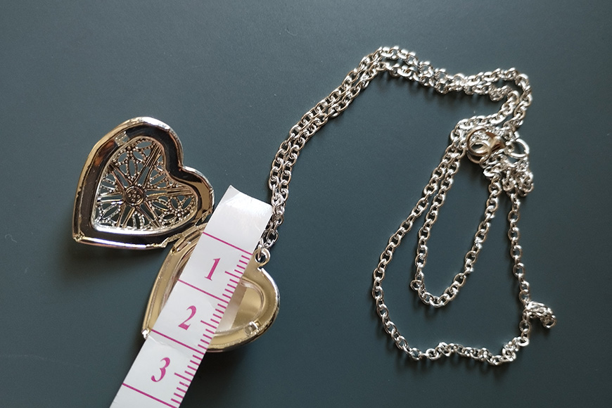 Heart-shaped locket with filigree design next to a measuring tape and silver chain on a dark surface.
