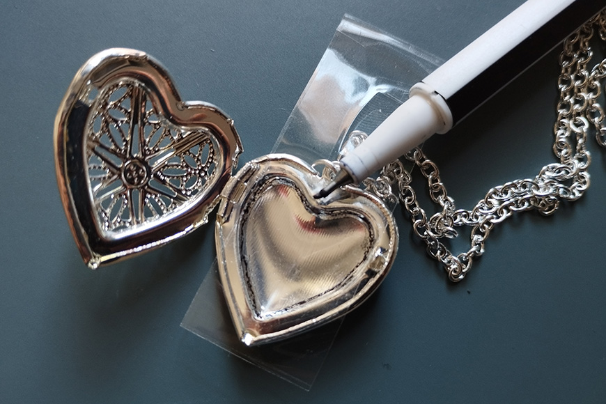 A locket in the shape of a heart with a compass design inside, next to a pen and a chain on a dark surface.