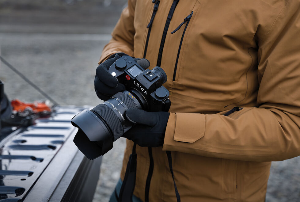 A person in a tan jacket holding a leica camera with a black lens.