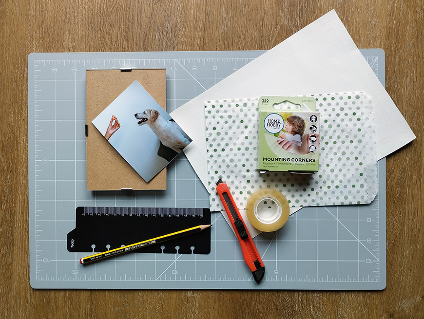 A selection of scrapbooking materials laid out on a cutting mat, including photographs, adhesive mounting corners, a ruler, a pen, and a roll of tape.