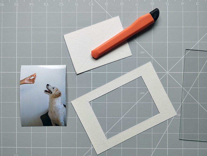 Crafting supplies including a photograph of a dog, a cutting mat, a utility knife, an envelope, and a paper frame.