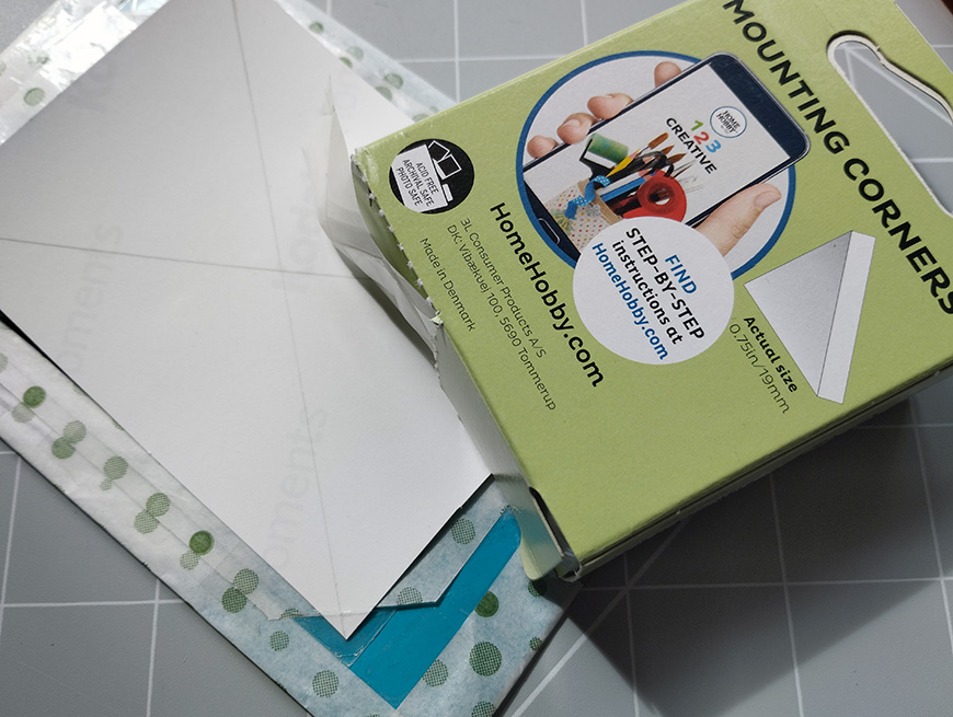 A box of mounting corners next to an envelope on a table with a cutting mat.