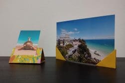 Two printed photographs propped up by simple paper holders on a desk, with one depicting a person enjoying the beach and the other showcasing a scenic coastal landscape.