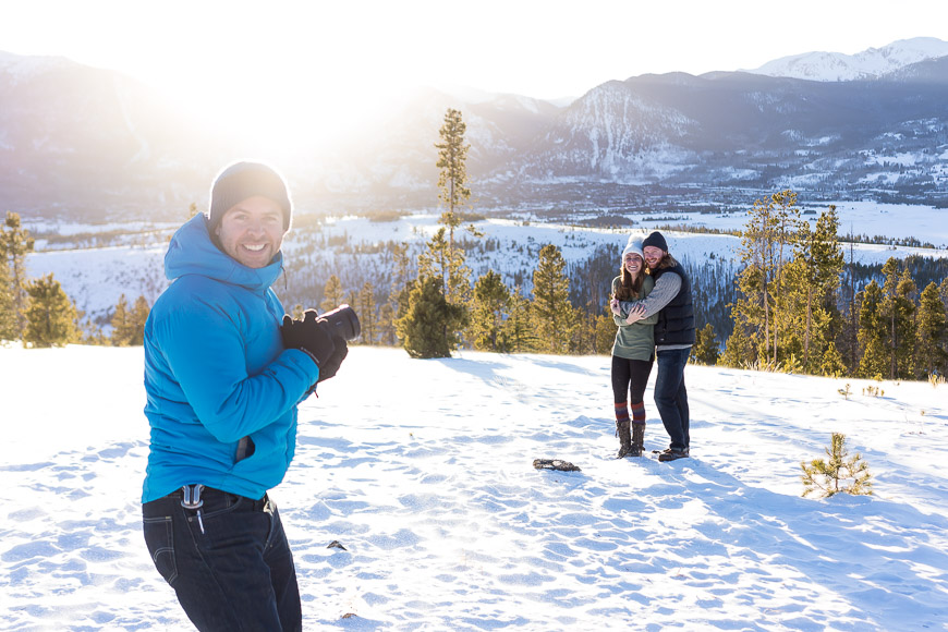 Man in a blue jacket taking a photo of a couple embracing in the snowy landscape at sunset.