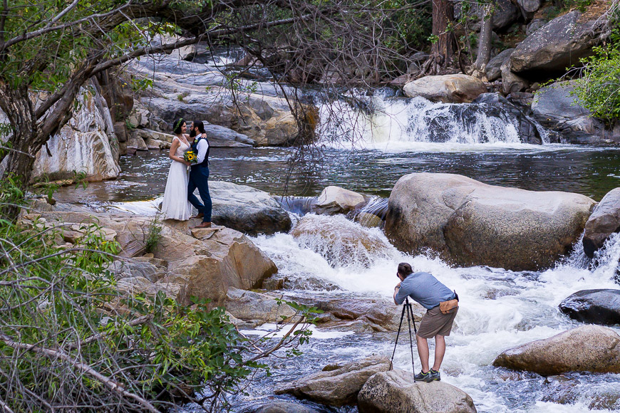 Photographer capturing a couple's wedding photos by a forest waterfall.