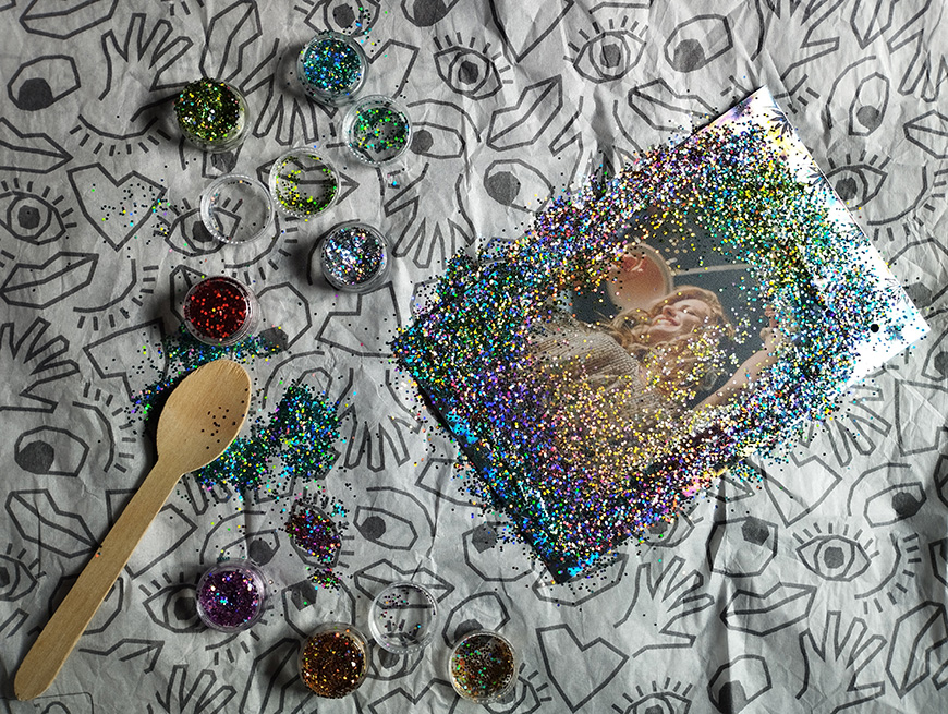 Craft glitter and sequins scattered on a tablecloth with artistic eye patterns, a wooden spoon, and a photograph partially covered by the colorful sparkles.