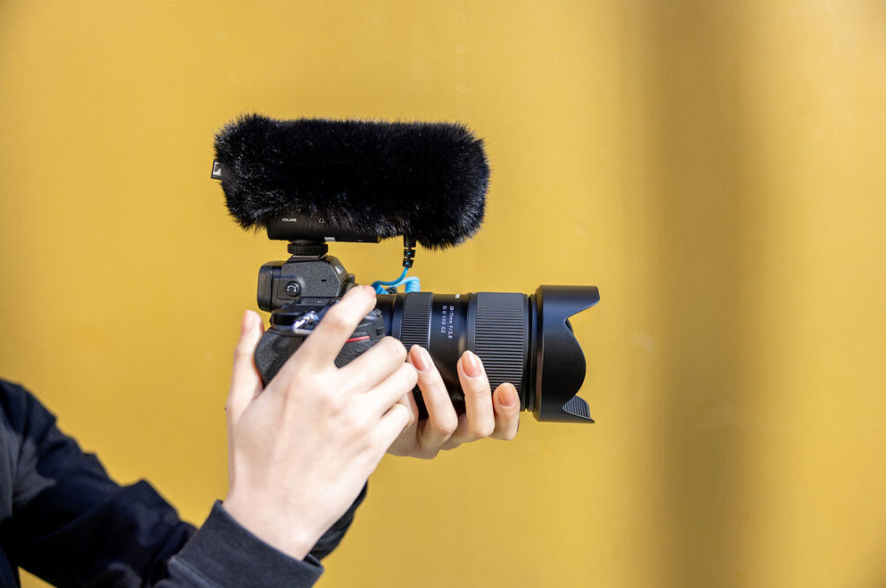 Person holding a dslr camera with an attached external microphone against a yellow background.