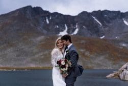 A couple in wedding attire embracing outdoors with a mountainous backdrop.
