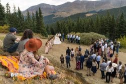 A scenic outdoor wedding ceremony with guests gathered around the bride and groom in a mountainous setting.