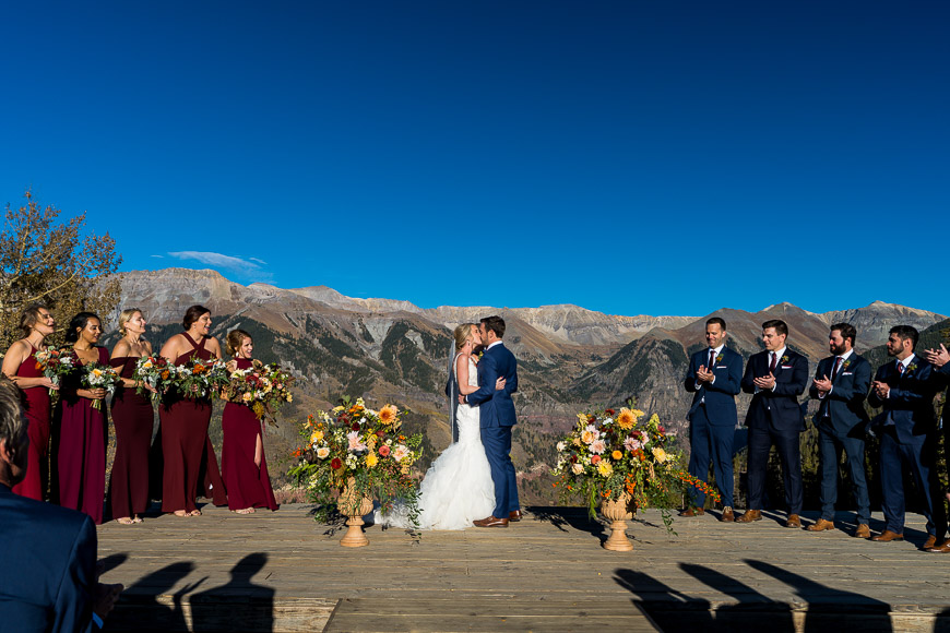 Wedding ceremony with a couple kissing on an outdoor altar, flanked by bridesmaids in burgundy dresses and groomsmen in blue suits, with a mountain backdrop under a clear blue sky.