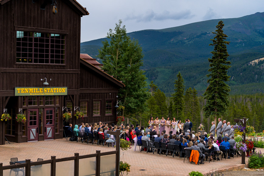 Outdoor wedding ceremony taking place in front of a rustic building with mountains in the background.