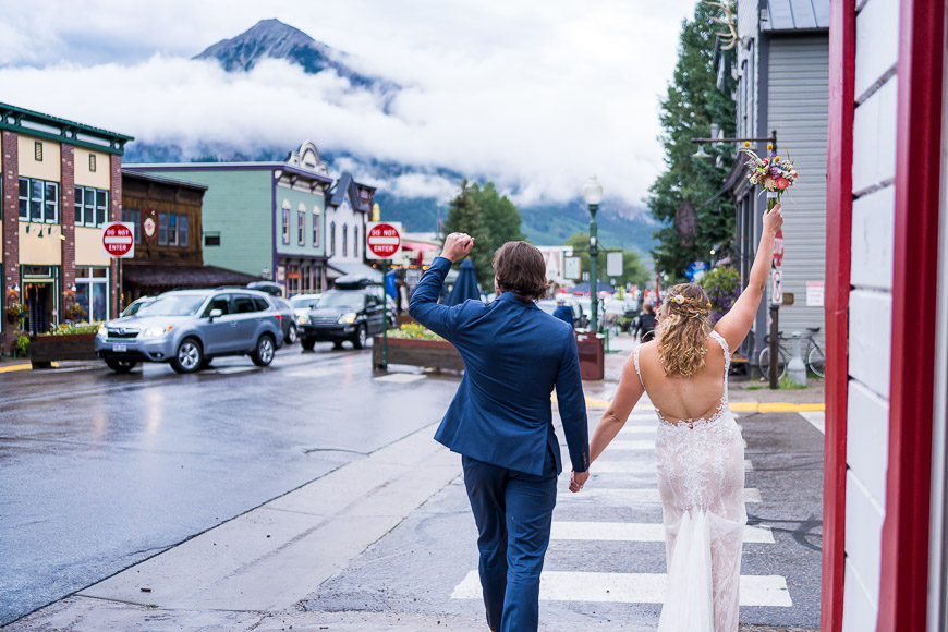 Wedding couple walking hand in hand on a town street with mountains partially obscured by clouds in the background.