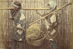 Two individuals in traditional attire carrying a large basket on a pole.