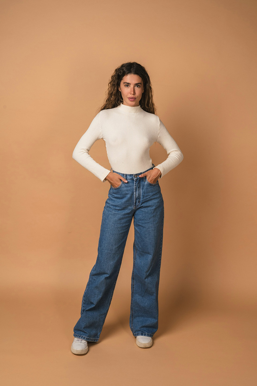 A person standing against a plain beige background, wearing a white long-sleeve top, blue high-waisted jeans, and white sneakers. Hands are on hips, and they are looking forward.
