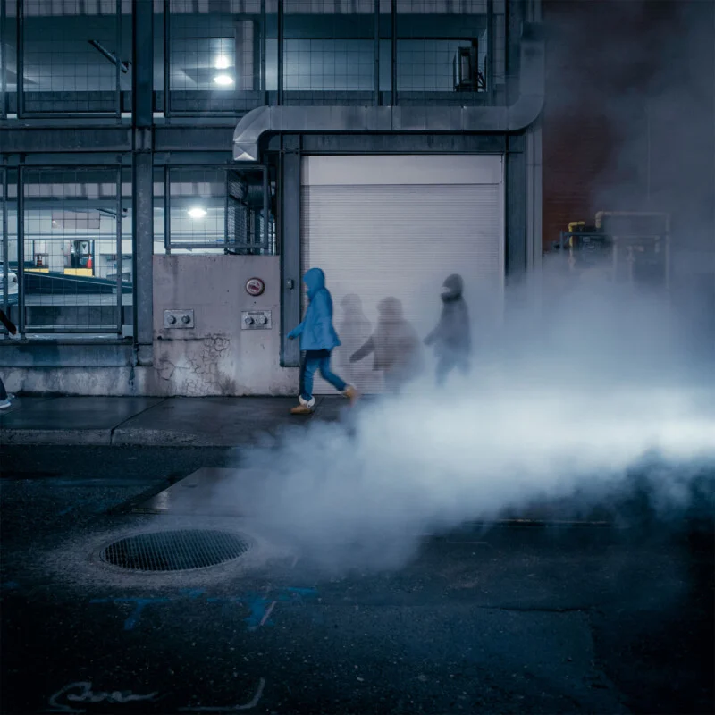 Individuals walking past a steam vent on a city street at night.