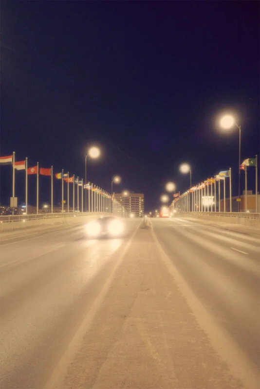 Night view of a well-lit, empty bridge adorned with flags, leading into a city skyline.