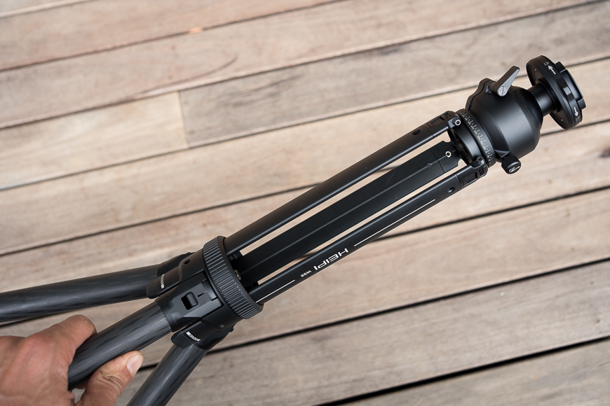 A person is holding a collapsed black tripod against a wooden plank background.