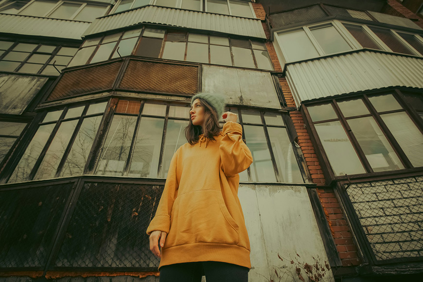 Person in a yellow hoodie and blue hat stands in front of a windowed, multi-story building, looking off to the side.